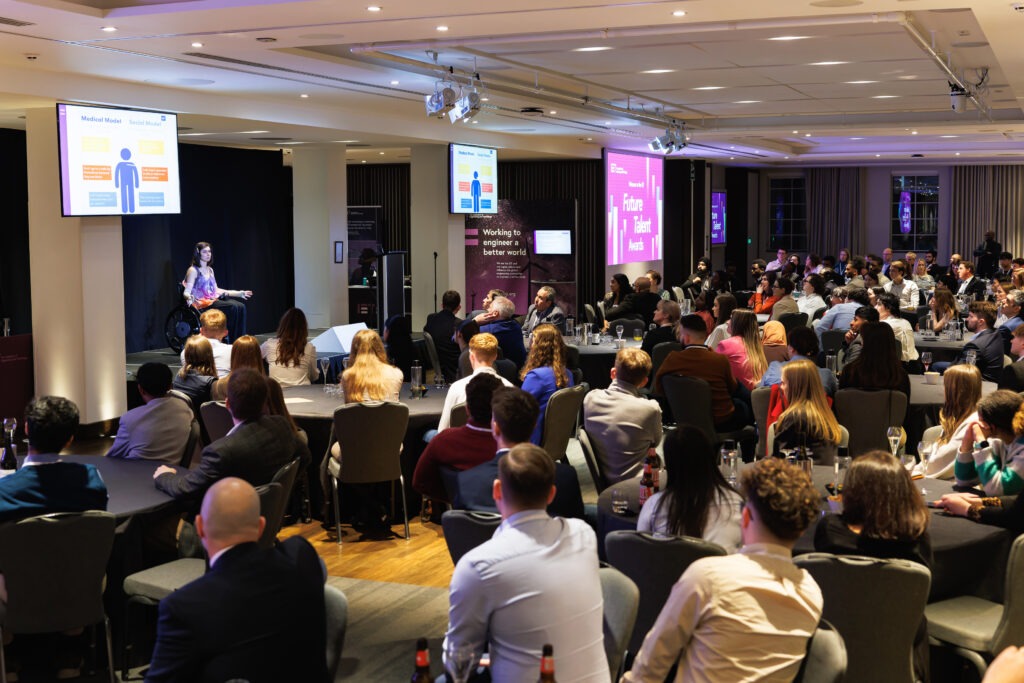 A large audience looking at a white dark haired middle aged woman on a stage with a screen depicting the difference between the health model of disability and the social model of disability