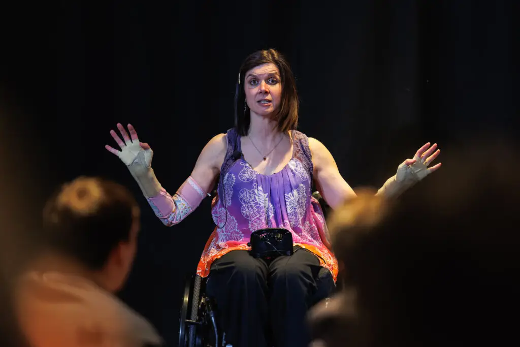 White dark haired middle aged woman, in a wheelchair. She is on stage, speaking to a large audience with her hands outstretched explaining about the social model of disability