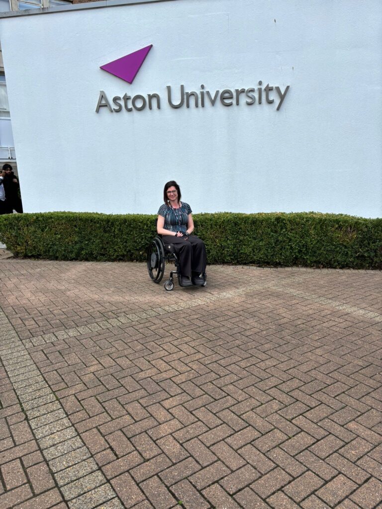 Katy sitting in a wheelchair under Aston University's sign